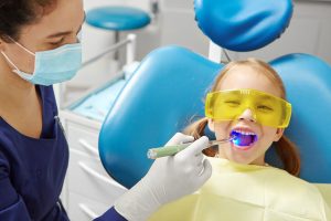 Process Of Drying The Dental Seal After Treatment Of The Patient's Tooth In Pediatric Dentistry. Little Girl In Dentist Office.